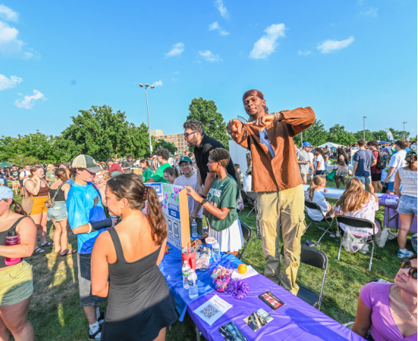 Students at an outdoor involvement fair at Michigan State University gather around a purple table filled with flyers, water bottles and decorations. A student on the right stands on a chair making a heart symbol with their hands, promoting engagement and campus pride under a clear blue sky.